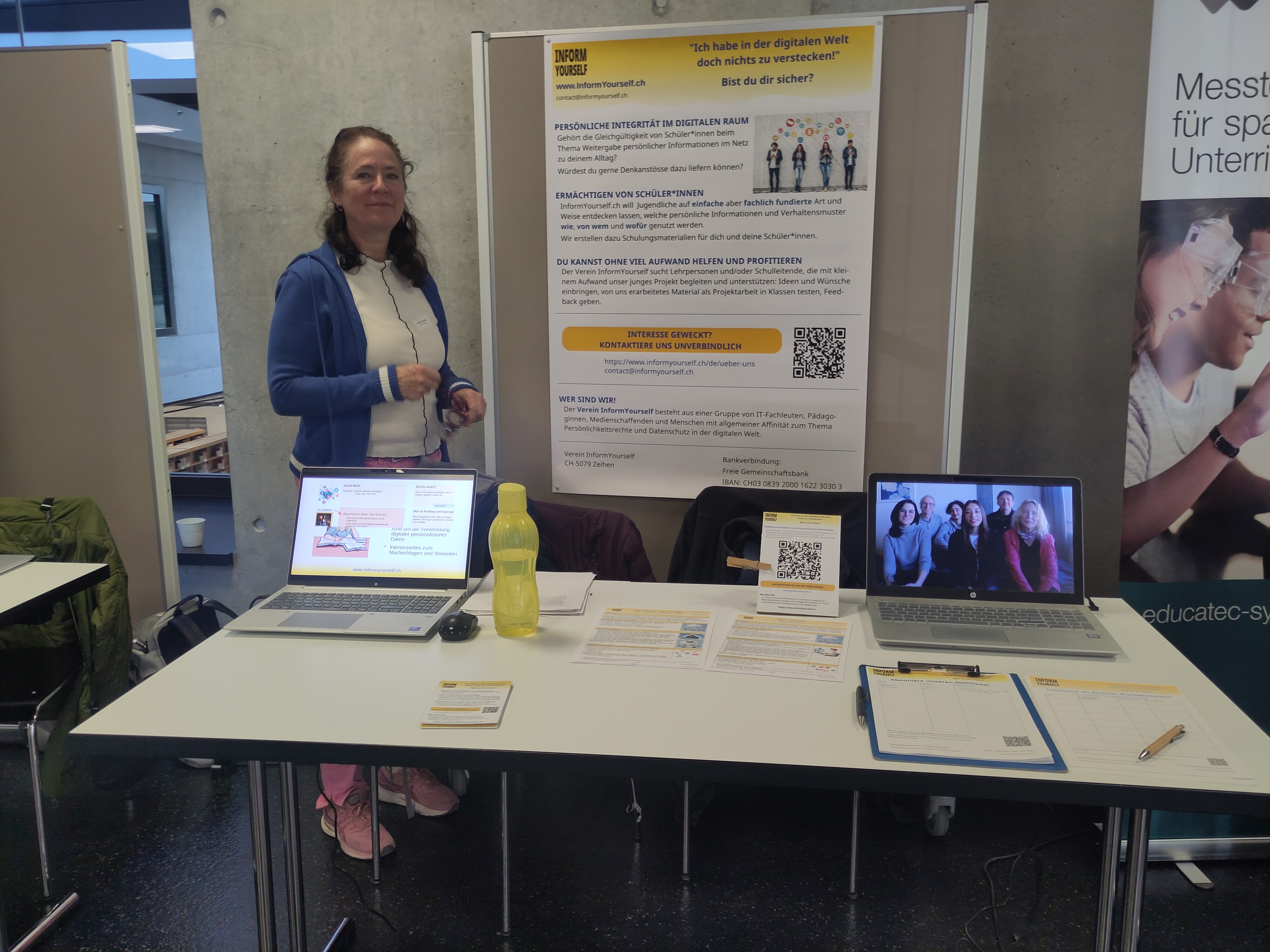 Nathalie von InformYourself steht neben dem Poster von InformYourself und hinter dem Tisch mit zwei Laptops und Informationsmaterial an unserem Stand anlässlich des Open Education Day in Bern